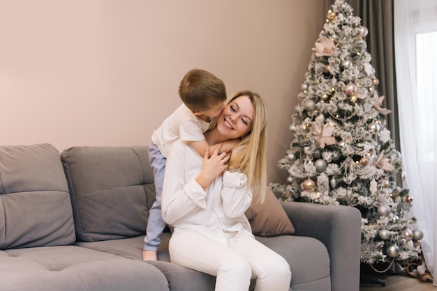 Mooie jonge moeder speelt en lacht met haar lachende zoontje thuis op de achtergrond van de kerstboom