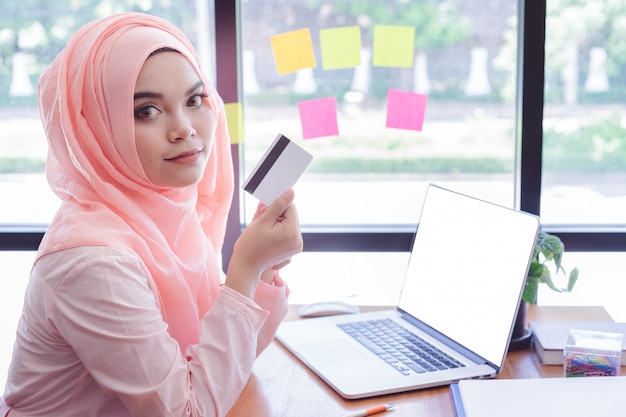 Mooie jonge moslimvrouw die een creditcard met laptop in bureau toont. Laptop met leeg scherm