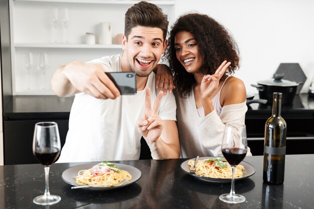 Mooie jonge multi-etnisch koppel met een romantisch diner thuis, rode wijn drinken en pasta eten, roosteren, een selfie nemen