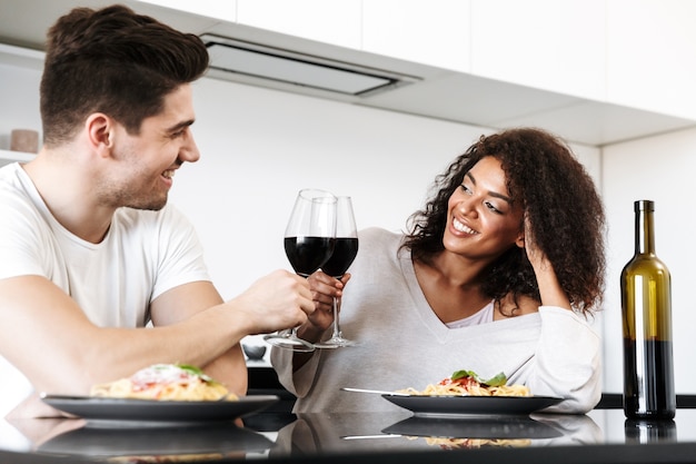 Mooie jonge multi-etnisch paar met een romantisch diner thuis, rode wijn drinken en pasta eten, roosteren