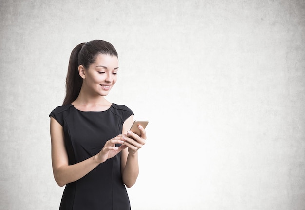 Mooie jonge onderneemster die in een zwarte kleding haar smartphonescherm bekijkt. Een betonnen muur achtergrond. Bespotten