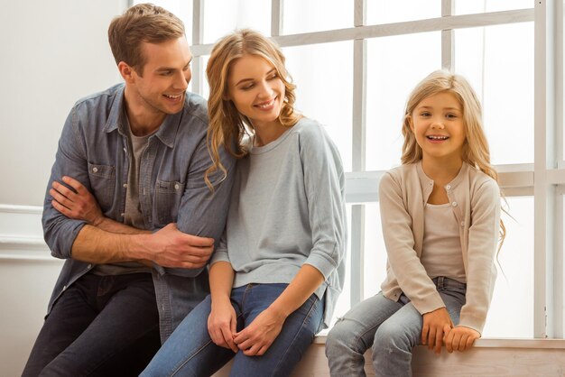 Mooie jonge ouders die naar hun mooie dochtertje kijken en glimlachen terwijl ze thuis bij het raam zitten