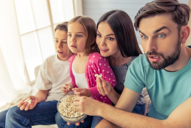 Mooie jonge ouders, hun dochter en zoon kijken tv, eten popcorn en laten een verrassing zien, terwijl ze thuis op de bank zitten