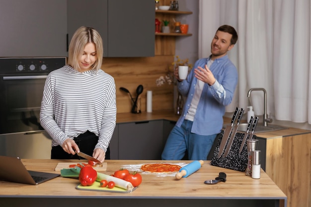 Mooie jonge paar is aan het praten en glimlachen tijdens het koken van gezond voedsel in de keuken thuis De man drinkt een kopje koffie en praat met de vrouw