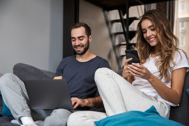 Mooie jonge paar ontspannen op een bank thuis, met behulp van laptop en mobiele telefoon