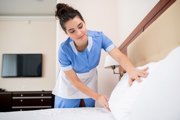 Mooie jonge professionele kamermeisje witte schone kussens aanbrengend terwijl bukken bed in hotelkamer