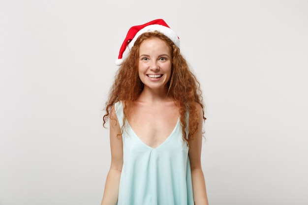 Mooie jonge roodharige Santa meisje in lichte kleding, kerstmuts geïsoleerd op een witte achtergrond, studio portret. Gelukkig Nieuwjaar 2020 viering vakantie concept. Bespotten kopie ruimte. Camera kijken.