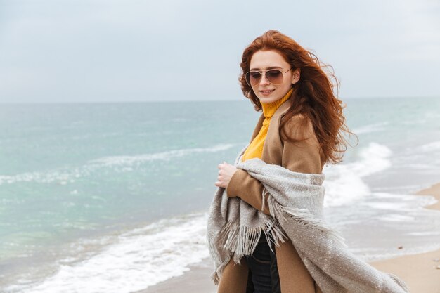 Mooie jonge roodharige vrouw met een herfstjas die op het strand loopt en deken vasthoudt