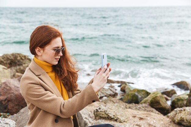 Mooie jonge roodharige vrouw met een herfstjas die op het strand loopt en een selfie maakt