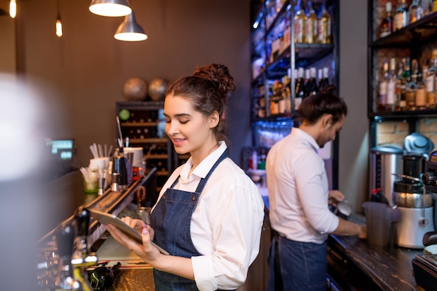 Mooie jonge serveerster met tablet scrollen door online bestellingen terwijl haar collega thee bereidt voor gasten op achtergrond