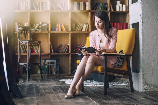 Mooie jonge slimme vrouw zit met het elektronische boek in de bibliotheek op een stoel