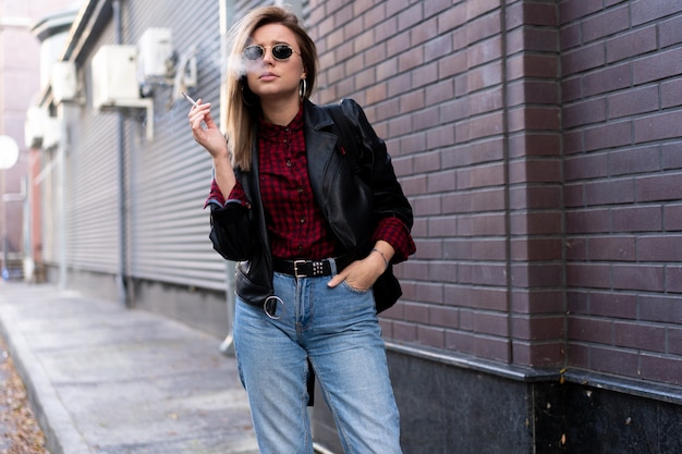 Mooie jonge stijlvol geklede blanke vrouw rookt sigaret op straat roken slechte gewoonte nicotineverslaving ongezonde levensstijl moderne vrouw leren jas en jeans lopen buiten zonnebril