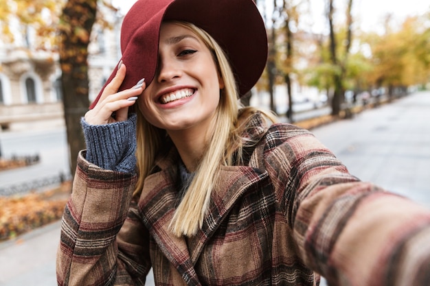 Mooie jonge stijlvolle blonde vrouw draagt een jas buiten wandelen, een selfie met mobiele telefoon nemen