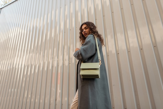 Mooie jonge stijlvolle modelvrouw met krullend haar in een modieuze groene jas met een dameshandtas loopt in de buurt van een metalen muur op straat. Vrouwelijke herfst casual stijl en mode