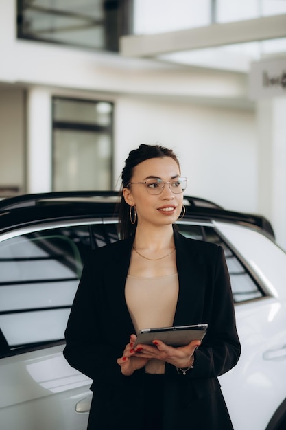 mooie jonge verkoopsvrouw in een elegant pak dealer die naast de auto staat bij een nieuwe moderne dealership