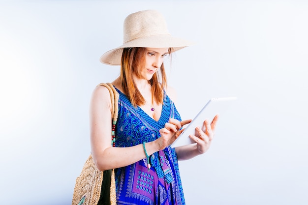 Mooie jonge volwassen vrouw in strand hoed en jurk kijken naar de status van reisreserveringen op de tablet op witte achtergrond. reisorganisatie concept. vlucht status