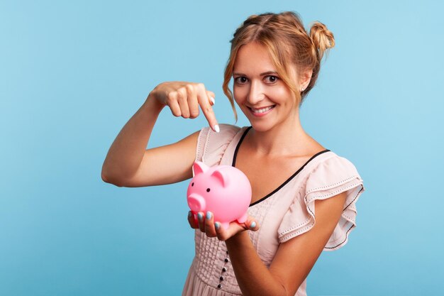 Mooie jonge volwassen vrouw in zomerjurk die een spaarvarken vasthoudt en met de wijsvinger wijst, wat suggereert dat je munten erin moet doen, geld besparen. Indoor studio opname geïsoleerd op blauwe achtergrond.