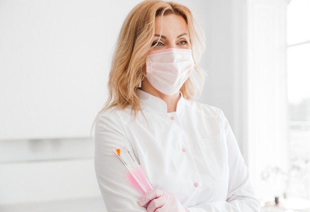 Mooie jonge vrouw arts schoonheidsspecialiste in een wit uniform en een beschermend masker op haar gezicht poseren met borstels in haar handen.