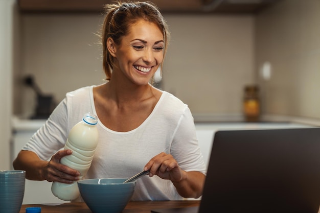 Mooie jonge vrouw bereidt haar gezond ontbijt in haar keuken en gebruikt laptop om een videochat met iemand te maken.