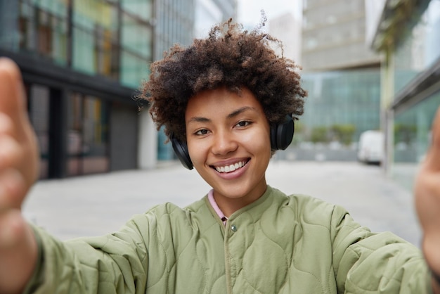 Mooie jonge vrouw brengt vrije tijd door in stedelijke omgeving maakt selfie foto's luistert muziek via draadloze koptelefoon draagt jas heeft goed humeur poses tegen wazige achtergrond Vrouwelijke reiziger in stad