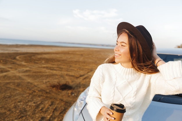 Mooie jonge vrouw buiten op het strand tijdens zonsondergang, zittend op een auto, koffie drinken