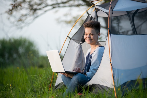 Mooie jonge vrouw die aan de camera glimlachen die vreugdevol in een tent zitten terwijl het kamperen gebruikend haar laptop