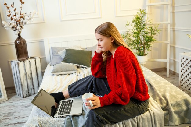 Mooie jonge vrouw die aan een laptop werkt die thuis op het bed zit, ze drinkt koffie en glimlacht. Thuiswerken tijdens quarantaine. Meisje in trui en spijkerbroek thuis op bed