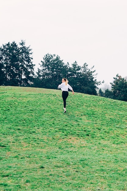 Mooie jonge vrouw die de groene heuvel oploopt in parkpijnbomen op het achtergrondvrijheidsconcept