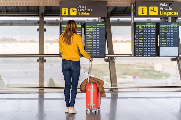 Mooie jonge vrouw die de vertrektijd van haar vlucht controleren op de informatieborden op de luchthaven met haar bagage