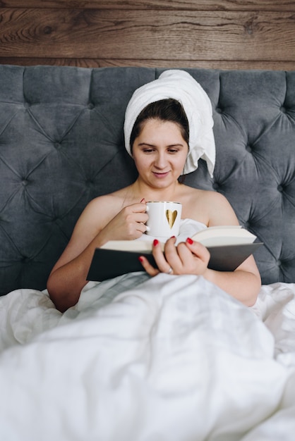 Mooie jonge vrouw die een boek in bed leest en een kop van koffie drinkt