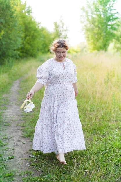 Mooie jonge vrouw die een elegante witte jurk draagt die met een glimlach op een weg in het bos staat met zonnestralen die door de bladeren van de bomen stralen