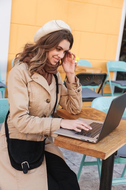 Mooie jonge vrouw die een jas draagt die laptopcomputer gebruikt tijdens het zitten bij de outdooors van het café