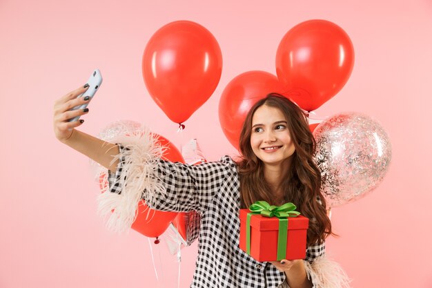 Mooie jonge vrouw die een jas draagt die zich geïsoleerd over roze achtergrond bevindt, viert, houdt bos van ballons vast, neemt een selfie