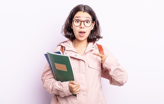 Mooie jonge vrouw die geschokt en verrast kijkt met wijd open mond, wijzend naar zichzelf. student met boeken concept