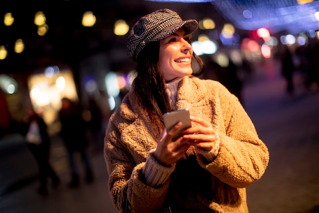 Mooie jonge vrouw die haar mobiele telefoon op straat gebruikt in de kersttijd