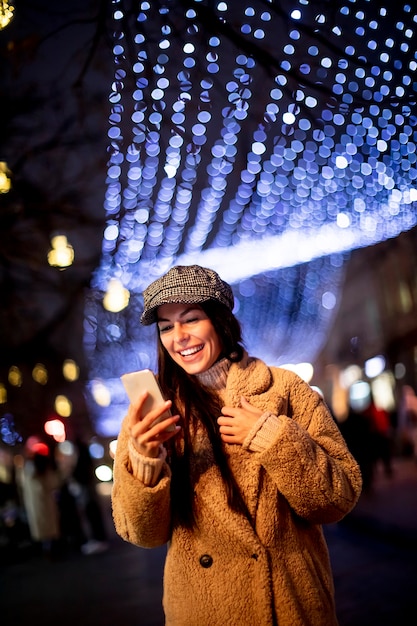 Mooie jonge vrouw die haar mobiele telefoon op straat gebruikt in de kersttijd