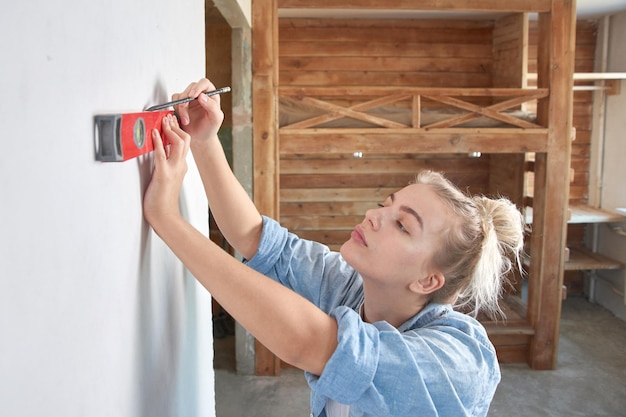 Mooie jonge vrouw die huisrenovatie doet, de uitlijning van de muur observeert met behulp van het waterniveau van het gebouw op een houten stapelbedachtergrond, DIY