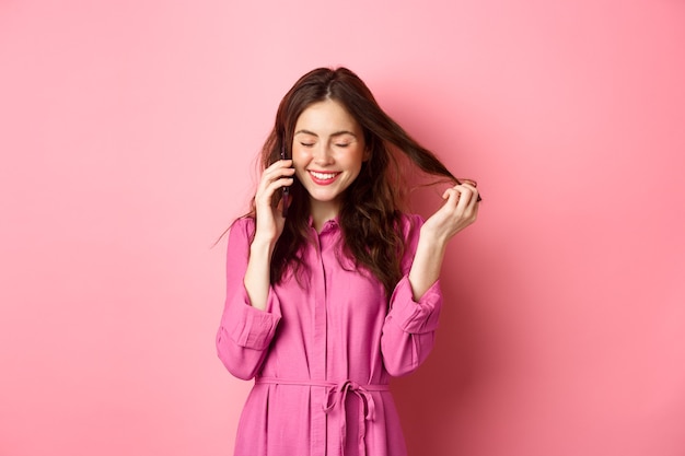 Mooie jonge vrouw die iemand belt, lacht en glimlacht tijdens telefoongesprek, praat met vriend en speelt met haarlokken, staande tegen roze muur.