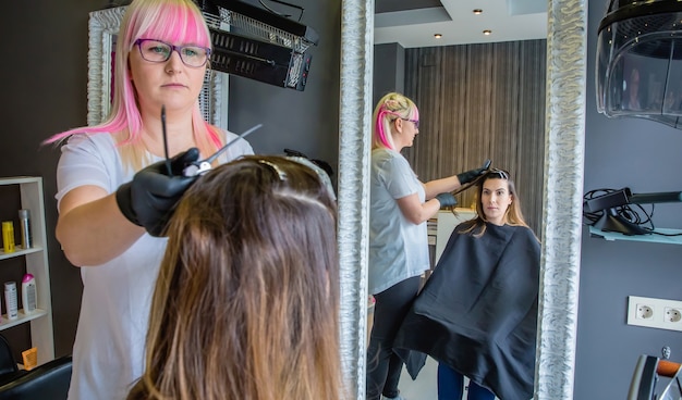 Mooie jonge vrouw die in een spiegel naar de kapper kijkt die het haar kamt tijdens het veranderen van haarkleur