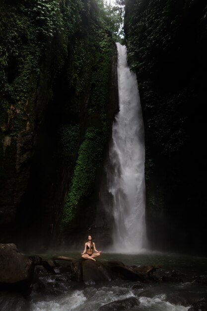 Mooie jonge vrouw die in lotusbloempositie mediteren terwijl het doen van yoga in een prachtig bos dichtbij waterval. Mooie vrouwelijke beoefenen van yoga op rots in de buurt van tropische waterval