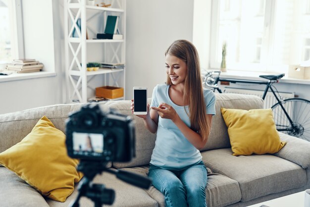 Mooie jonge vrouw die lacht en kopieert op haar smartphone terwijl ze thuis een video op sociale media maakt