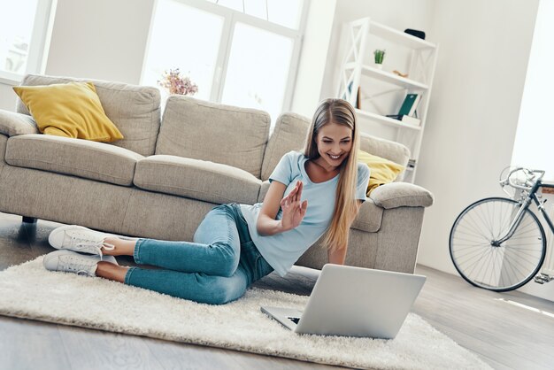 Mooie jonge vrouw die met glimlach zwaait en laptop gebruikt terwijl ze thuis rust