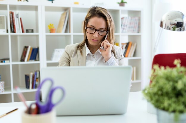 Mooie jonge vrouw die met haar mobiele telefoon werkt in haar kantoor.