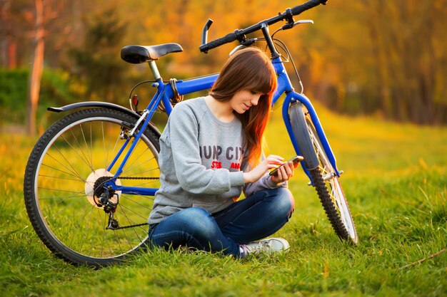 Mooie jonge vrouw die mobiele telefoonzitting naast haar fiets gebruiken bij zonsondergang