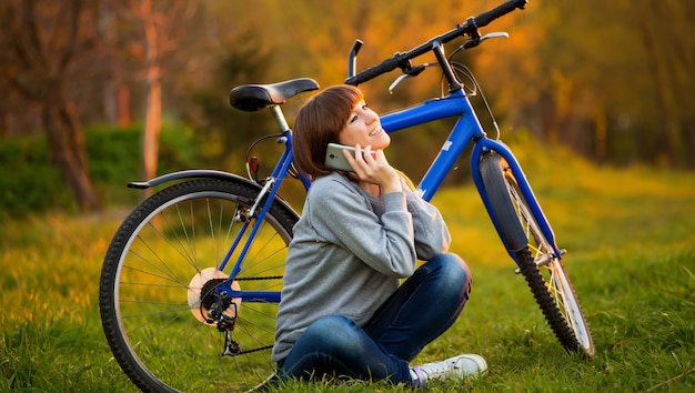 Mooie jonge vrouw die mobiele telefoonzitting naast haar fiets gebruiken bij zonsondergang