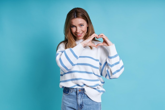 Mooie jonge vrouw die naar de camera kijkt en haar duim laat zien terwijl ze tegen een blauwe achtergrond staat