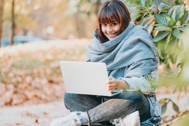 Mooie jonge vrouw die op de bank in een park zit en op een laptop werkt terwijl ze plezier heeft met het nemen van