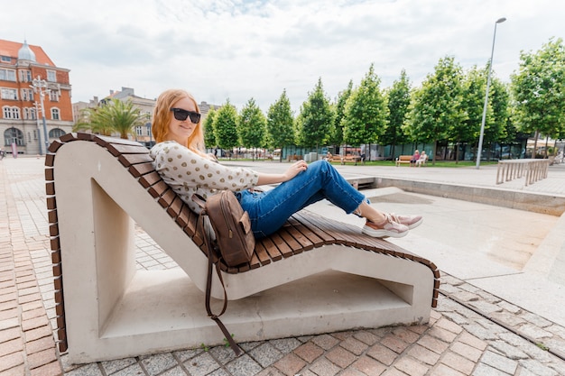 Mooie jonge vrouw die op een bank bij de backgroung van de stad rust