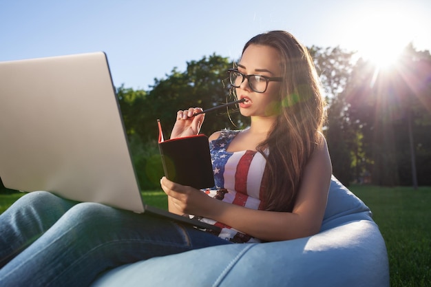 Mooie jonge vrouw die op een zitzak zit, gebruikt een laptop terwijl ze op het gras in het park op de zon rust