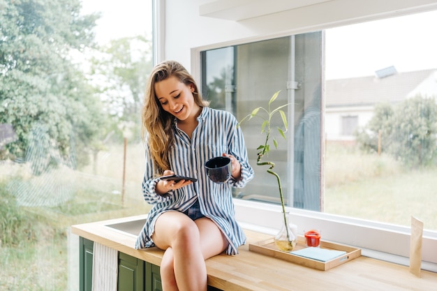 mooie jonge vrouw die op het aanrecht zit en op de mobiele telefoon bladert in de moderne keuken